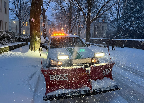 Einsatzfahrzeug mit Schneeschieber räumt abends bei Schneefall die Straße.